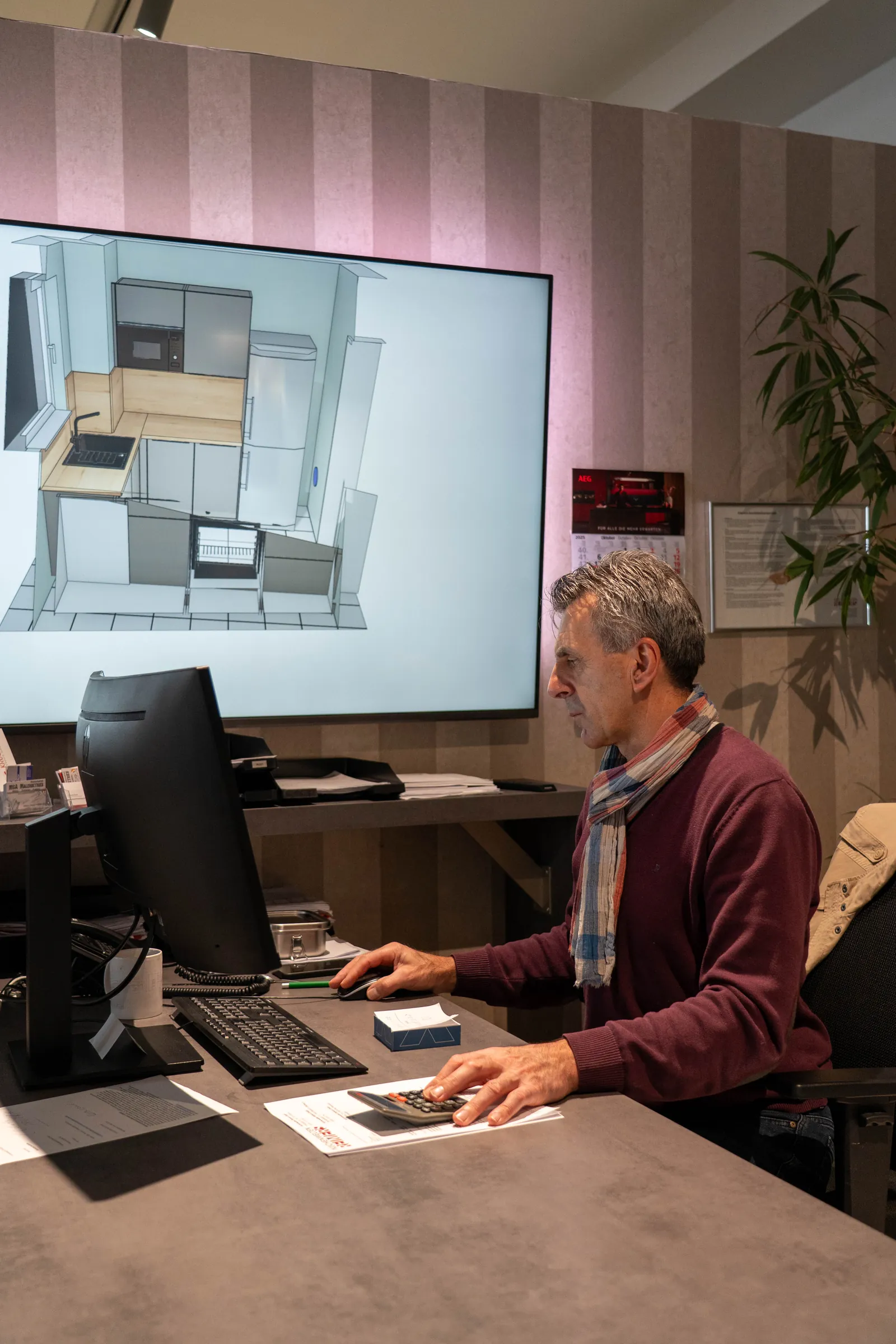 Küchenplaner am Arbeitsplatz, auf dem Großbildschirm eine 3D-Planung der neuen Küche