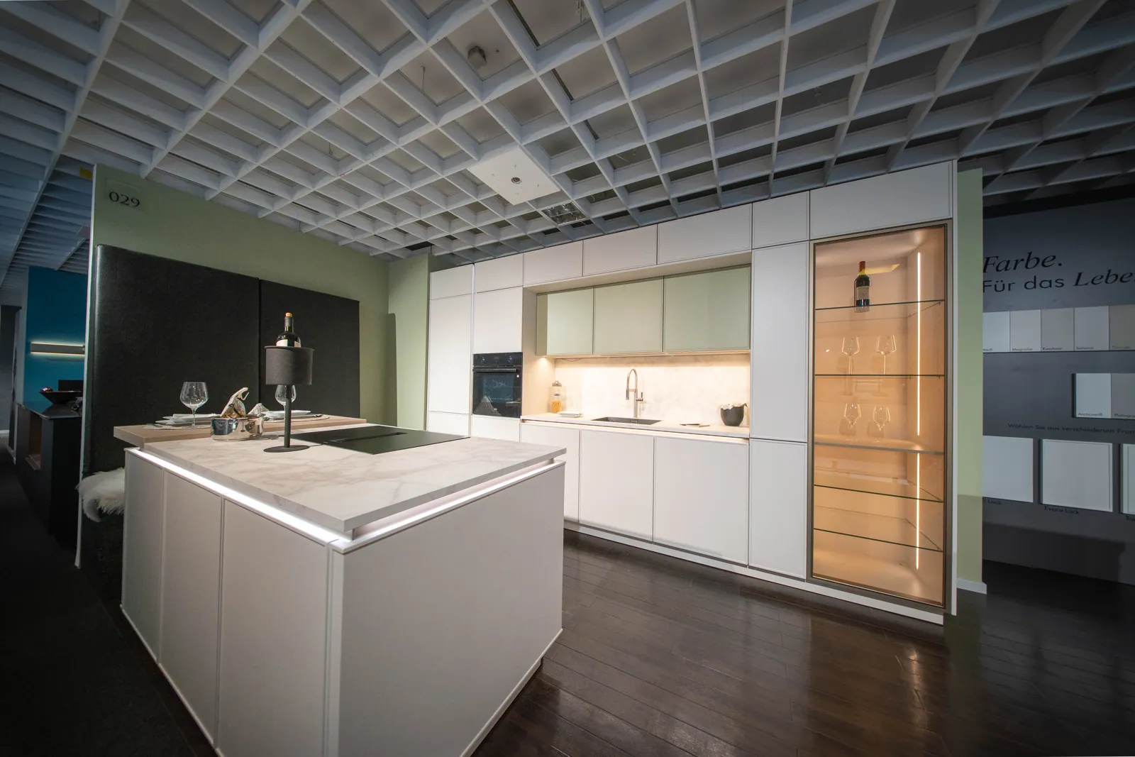 Weiße Kücheninsel und Hochschrankwand mit salbeigrünen Oberschränken in der Ausstellung Bonn